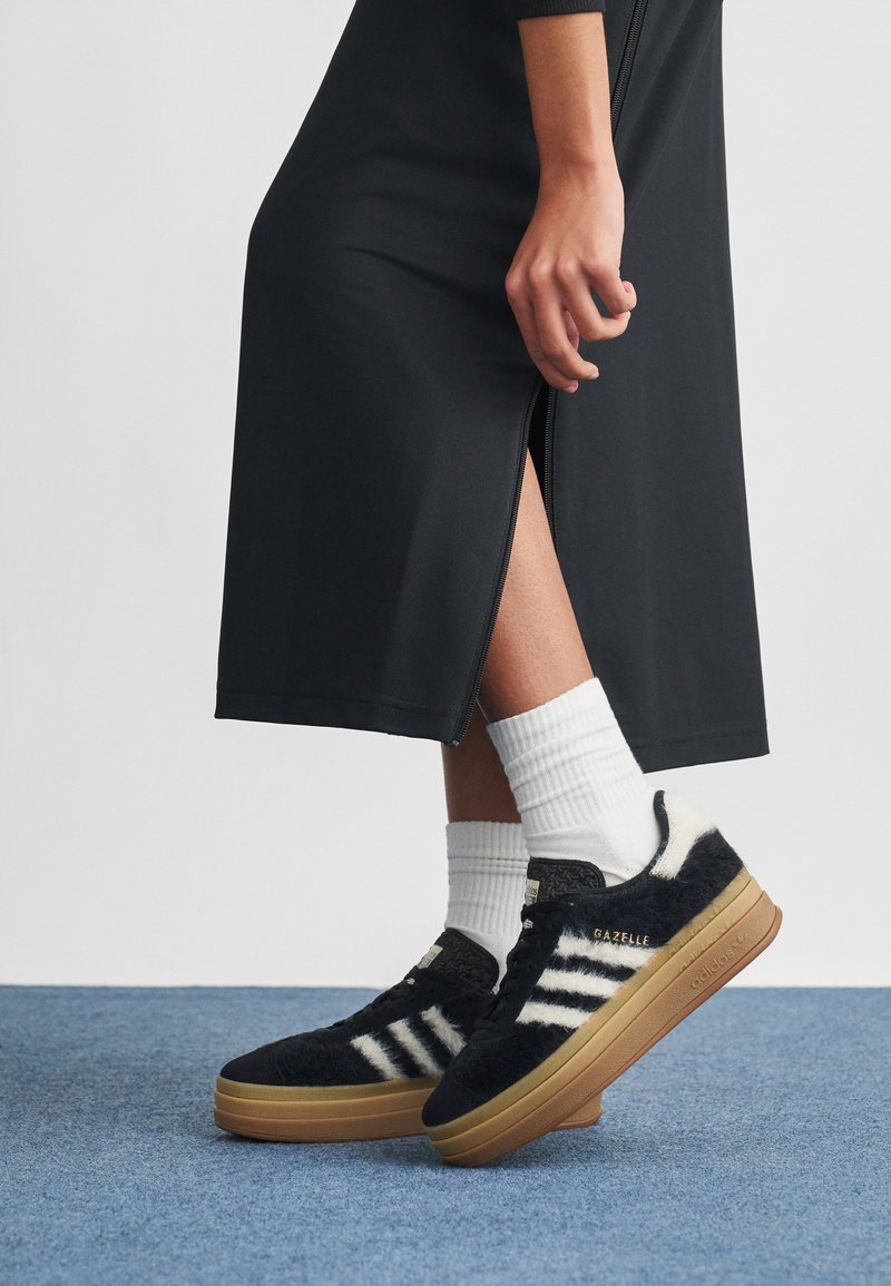 Sapatilhas Adidas Gazelle pretas felpudas com três riscas brancas, sola de borracha castanha e parte superior texturada. Usadas com meias brancas até ao tornozelo sobre um tapete azul.