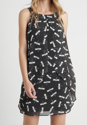 Sleeveless black dress with white dragonfly print worn by a standing person with long blonde hair, hand resting near dress hem.