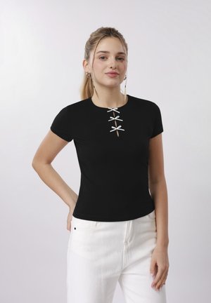 Young woman with blonde hair tied back wears a black short-sleeve top with three silver bows and white pants, standing against a plain background.