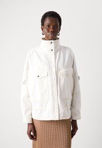 White denim jacket with a high collar, two front pockets, and snap buttons. Features contrasting stitching and a relaxed fit.