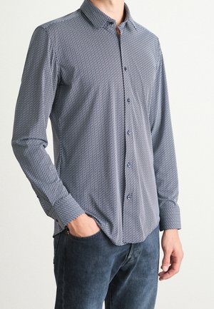 Mann trägt ein langärmeliges Hemd mit kleinem weißem geometrischem Muster und dunkle Jeans, Hand in der Tasche, vor schlichtem Hintergrund.
