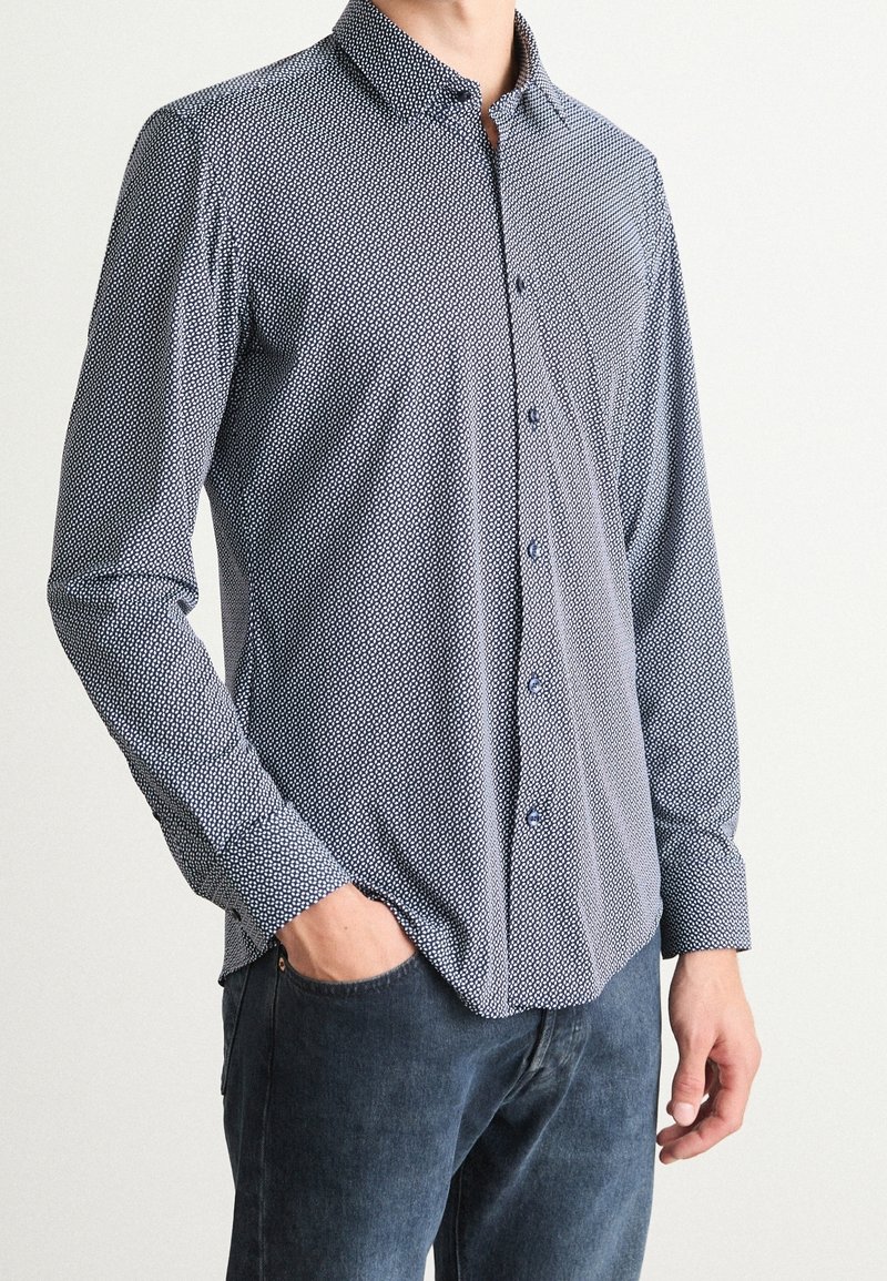 Homme portant une chemise à manches longues avec un petit motif géométrique blanc et un jean foncé, main dans la poche, sur un fond uni.