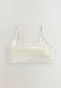 Reggiseno color crema con sottili spalline regolabili, design senza cuciture e un'etichetta che presenta tre contorni a forma di cuore nella parte inferiore anteriore.