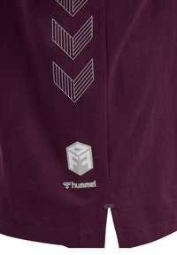 Maron t-shirt har et struktureret stof, et grafisk mønster af chevroner i hvid, og kontrasterende logo accenter ved bunden.