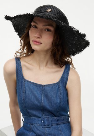 Femme aux cheveux bruns bouclés portant un chapeau de paille noir à large bord et une robe en denim sans manches ceinturée, sur fond blanc.