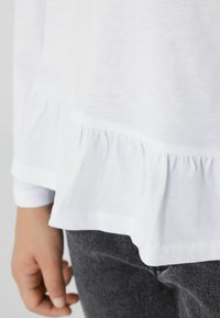 Biała bawełniana bluzka z falbaną na dole. Rękawy mają wykończenie w mankiet, zestawione z szarymi teksturowanymi spodniami, ukazując zbliżenie na materiał.