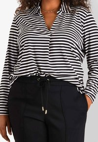 Schwarz-weiße gestreifte Langarmbluse mit Kragen, in entspanntem Schnitt. Kombiniert mit schwarzen Jogginghosen mit Kordelzug und goldenen Hardware-Details.