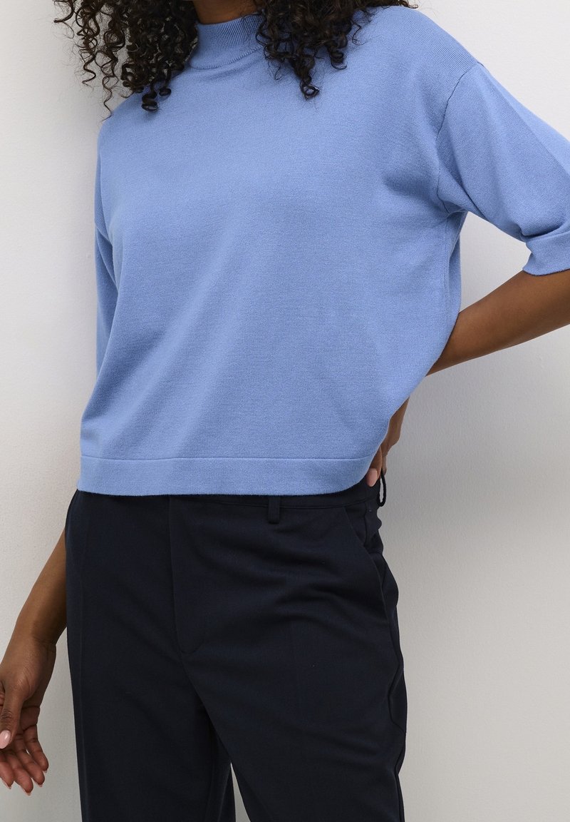 Femme portant un pull léger bleu clair à manches courtes légèrement rentré dans un pantalon noir taille haute, debout avec une main sur la hanche.