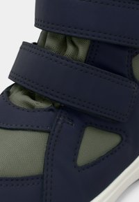 Navy modré sneakersy s olivově zelenými síťovanými panely, vybavené dvěma suchými zipy a texturovanými detaily na syntetickém materiálu. Bílá gumová podrážka.