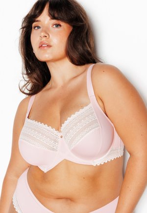 Femme à la carnation moyenne portant un soutien-gorge en dentelle rose clair et des sous-vêtements assortis sur fond blanc.