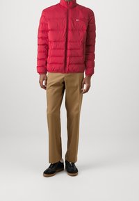 Homme portant une veste matelassée rouge, un pantalon beige à jambe droite et des baskets noires avec des semelles brunes, se tenant devant un fond uni.