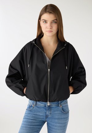 Giovane donna con giacca nera con cappuccio e zip e jeans blu, in piedi con le mani in tasca contro uno sfondo bianco.