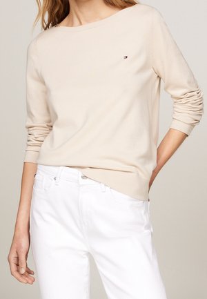 Femme portant un pull beige à manches longues avec un petit logo sur la poitrine et un pantalon blanc, debout avec une main dans la poche.
