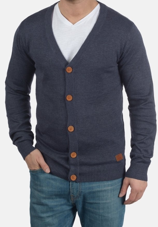 Homme portant un cardigan bleu foncé boutonné par-dessus une chemise blanche à col en V et un jean bleu, mains dans les poches, torse visible.