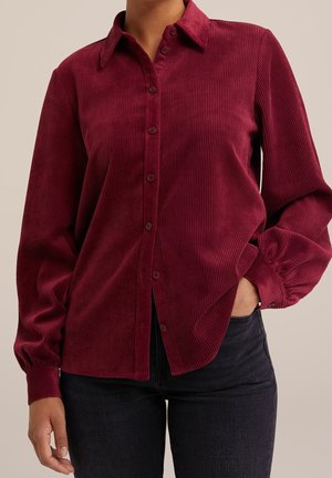 Vrouw draagt een bordeauxrode lange mouwen ribfluwelen blouse met knopen, één hand in de zak van een zwarte spijkerbroek, tegen een effen beige achtergrond.