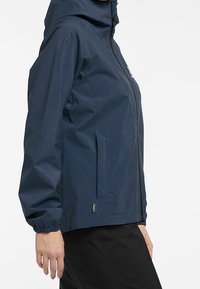 Veste imperméable marine avec capuche, fermeture éclair frontale, poches latérales, poignets élastiques et texture lisse et légère.