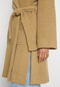 Cappotto in pile color cammello con finitura strutturata, vita con cintura, maniche ampie e spacchi laterali, abbinato a denim chiaro.