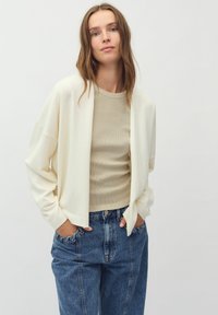 Cardigan lejer de culoare crem deschis, cu mâneci largi, purtat cu un top bej ajustat și blugi albaștri cu talie înaltă.