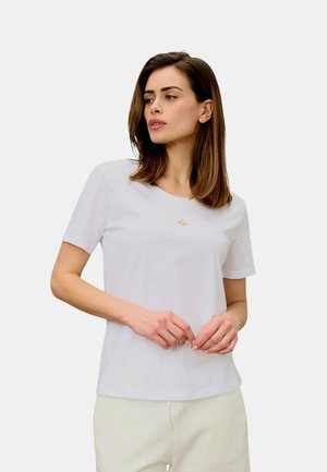 Donna con capelli castani fino alle spalle, che indossa una semplice maglietta bianca a maniche corte e pantaloni chiari, guarda di lato.