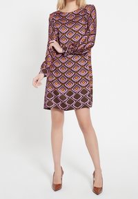 Modellen poserar med ena handen på höften, iförd en brun klänning med ett färgglatt geometriskt mönster och bruna högklackade skor framför en enkel bakgrund.