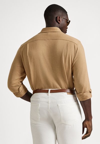 Vyras, dėvintis susukta smėlio spalvos ilgomis rankovėmis marškinius, baltas kelnes, rudos spalvos diržą ir akinius nuo saulės, žiūrimas iš nugaros priešais vienspalvį foną.