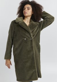 Faux fur olivengrøn frakke med dobbeltknaplukning, stor klapkrave og sidelommer, der fremviser et blødt, tekstureret stof.