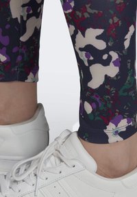 Ben som bär flerfärgade blommönstrade leggings och vita Adidas-sneakers med snören mot en enkel ljus bakgrund.