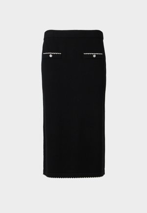 Femme aux cheveux courts et foncés portant un top noir à manches courtes et une jupe midi assortie avec un bord blanc et des détails de boutons, se tenant devant un fond blanc.