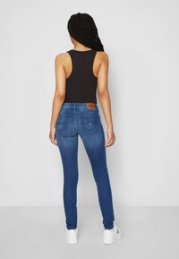 Jeans ajustados azules con una cinturilla oscura, dos bolsillos traseros y una etiqueta de marca marrón, combinados con una camiseta sin mangas negra y zapatillas blancas.