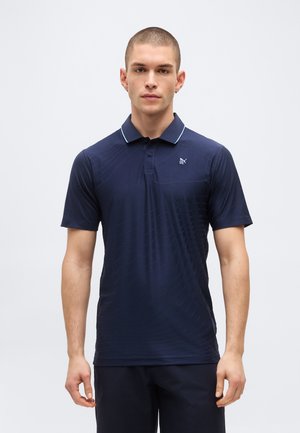 Νέος άνδρας με κοντά μαλλιά που φοράει ένα μπλε ναυτικό πόλο της Puma με διακριτικά σχέδια, στέκεται μπροστά σε ένα απλό φωτεινό φόντο.