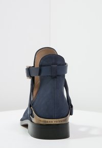 Botín de cuero azul marino con correa y hebilla, placa de tacón de bronce grabada "Rossetti Patented" y suela negra en capas.