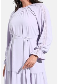 Vestido de lavanda claro con escote plisado, cinturón ajustable y detalles de encaje intrincados en las mangas. Faldón acampanado y textura suave.