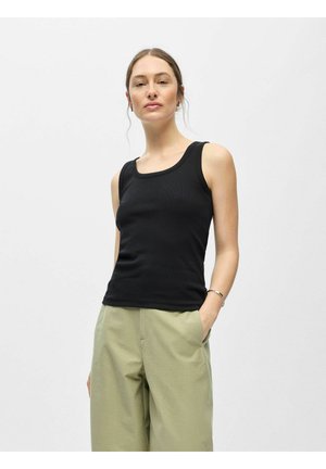 Femme portant un haut noir sans manches et un pantalon vert clair, debout avec une main dans la poche devant un fond gris uni.