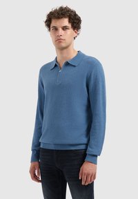 Blauwe fleecetrekker met een ritskraag, textuurstof, geribbelde zoom en manchetten, gedragen over een donkere spijkerbroek, model staat tegen een neutrale achtergrond.