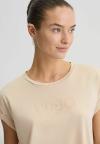 Camicia beige a maniche corte con colletto rotondo, caratterizzata da un logo testurizzato "LIU•JO 1995" di una tonalità leggermente più scura. Tessuto morbido, design minimal.