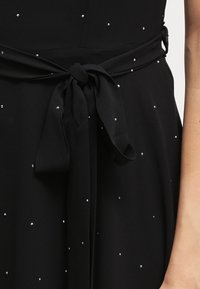 Musta mekko, jossa on hopeisia koristeluja ripoteltuna, vyö solmittuna vyötäröllä. Kangas on sileäpintainen ja laskeutuva.