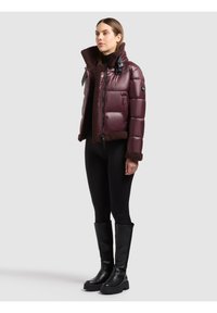 Femme portant une veste matelassée brun foncé brillante avec une bordure en peau lainée, des leggings noirs et des bottes noires montantes, debout devant un fond uni.