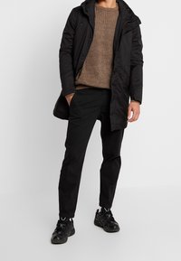 Man i brun stickad tröja, svart vadderad jacka, svarta byxor och svarta sneakers som står med en hand i fickan.