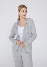 Světlá šedá mikina na zip vyrobená z měkkého materiálu, vybavená kapucí, předními kapsami a volným střih. Doplněno o odpovídající jogger kalhoty.