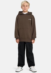 Barn med kort, blondt hår står i en brun hettegenser, svarte cargo-bukser og hvite sneakers, og poserer selvsikkert foran en ensfarget bakgrunn.
