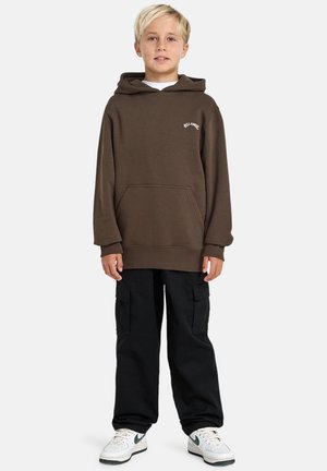 Un bambino con capelli biondi corti indossa un hoodie marrone, pantaloni cargo neri e sneaker bianche, posando con sicurezza su uno sfondo semplice.