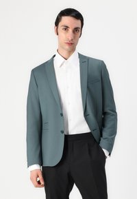 Blazer teal in tessuto liscio con bottoni singoli, abbinato a una camicia bianca con colletto e pantaloni neri. Design minimalista con tasche.