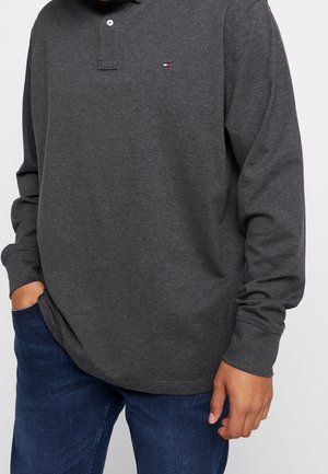 Polo de manga larga gris oscuro con tapeta abotonada, con un pequeño logo en el pecho izquierdo. Se lleva con vaqueros azules.