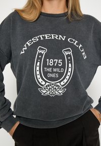 Pelēks sporta krekls ar baltu zirgskāpes grafiku, tekstu "WESTERN CLUB," "1875," un "THE WILD ONES," ar ribotām piedurknēm un apakšmalu.
