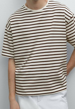 Personne portant un t-shirt blanc à manches courtes avec de fines rayures horizontales marron et un pantalon blanc, debout avec un bras plié derrière.