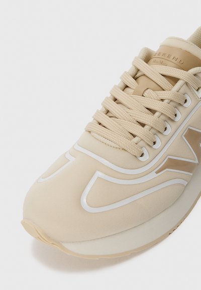 Beez sneaker paksude nööridega ja valgete aktsentjoontega varvaste ja külgede peal, näidates ülevalt-eest poolt detaili valge tausta ees.