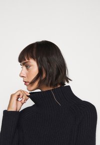 Pull en maille côtelée bleu marine avec une finition texturée, doté d'un col montant et d'un subtil accent doré au niveau du col.