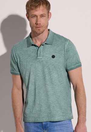 Homme blond portant un polo vert à manches courtes et un jean bleu, debout devant un fond clair avec son ombre visible.