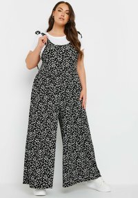 Svart blommig jumpsuit med vita blommor, design med vida ben, justerbara knytband, bärs över en vit t-shirt, ihop med vita sneakers.