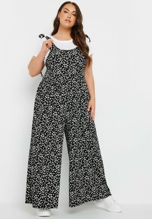 Sort blomsterjumpsuit med hvide blomster, vidde ben-design, justerbare bindebånd, båret over en hvid t-shirt, sat sammen med hvide sneakers.